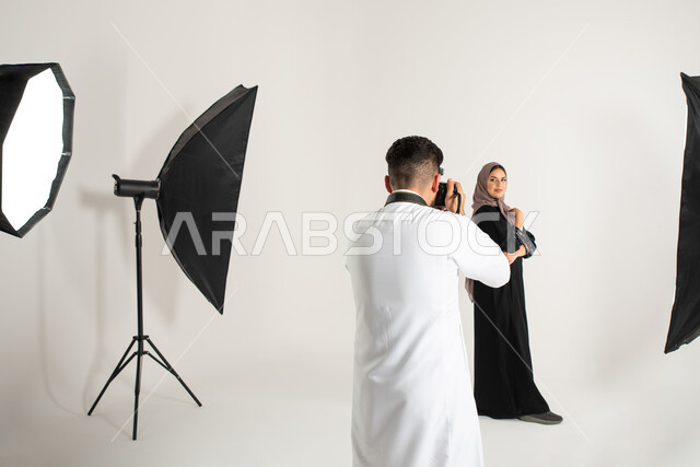 امرأة ترتدي عباءة نسائية خليجية، كاميرات ومعدات متطورة، بورتريه لجلسة تصوير في الاستوديو، مصور محترف عربي سعودي خليجي يقوم بالتقاط صور فوتوغرافية لعارضة أزياء وموديلات عصرية، تصوير اعلاني تسويقي، خلف الكواليس، خلفية بيضاء