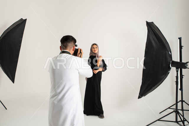 عارضة أزياء وموديلات عصرية، بورتريه لجلسة تصوير في الاستوديو، مصور محترف عربي سعودي خليجي يقوم بالتقاط صور فوتوغرافية، امرأة ترتدي عباءة نسائية خليجية، كاميرات ومعدات متطورة، تصوير اعلاني تسويقي، خلف الكواليس، خلفية بيضاء