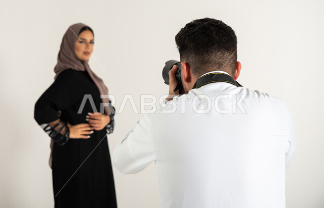 بورتريه لجلسة تصوير في الاستوديو، مصور محترف عربي سعودي خليجي يقوم بالتقاط صور فوتوغرافية لعارضة أزياء وموديلات عصرية، امرأة ترتدي عباءة نسائية خليجية، كاميرات ومعدات متطورة، تصوير اعلاني تسويقي، خلف الكواليس، خلفية بيضاء