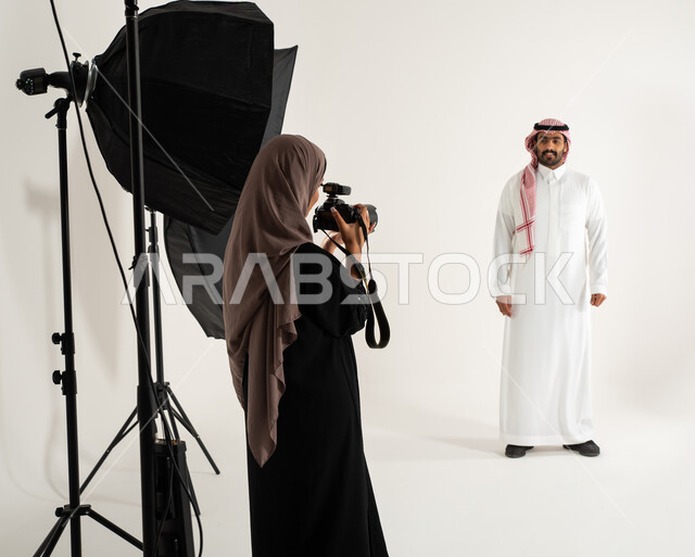 رجل يرتدي الزي السعودي التقليدي، تصوير اعلاني تسويقي، مصورة محترفة عربية سعودية خليجية تقوم بالتقاط صور فوتوغرافية، بورتريه لجلسة تصوير في الاستوديو، كاميرات ومعدات متطورة، خلف الكواليس، خلفية بيضاء