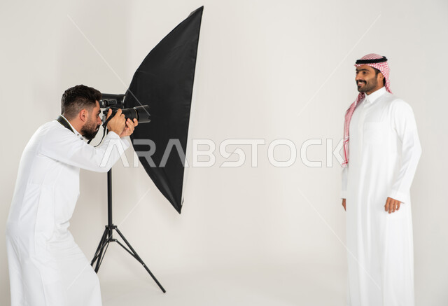 مصور محترف عربي سعودي خليجي يقوم بالتقاط صور فوتوغرافية لرجل يرتدي الزي السعودي التقليدي، تصوير اعلاني تسويقي، خلف الكواليس، بورتريه لجلسة تصوير في الاستوديو، كاميرات ومعدات متطورة، خلفية بيضاء