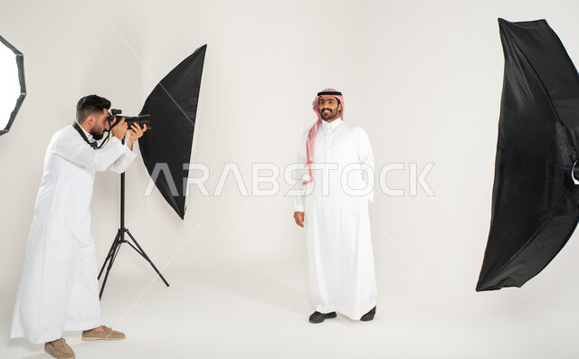 بورتريه لجلسة تصوير في الاستوديو، تصوير اعلاني تسويقي، كاميرات ومعدات متطورة، مصور محترف عربي سعودي خليجي يقوم بالتقاط صور فوتوغرافية لرجل يرتدي الزي السعودي التقليدي، خلف الكواليس، خلفية بيضاء