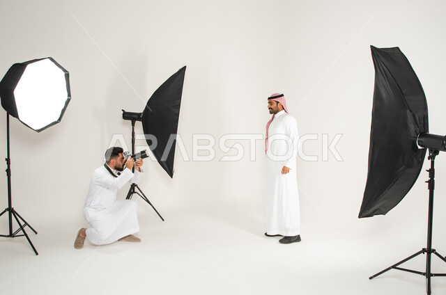 بورتريه لجلسة تصوير في الاستوديو، خلف الكواليس، كاميرات ومعدات متطورة، مصور محترف عربي سعودي خليجي يقوم بالتقاط صور فوتوغرافية لرجل يرتدي الزي السعودي التقليدي، تصوير اعلاني تسويقي، خلفية بيضاء