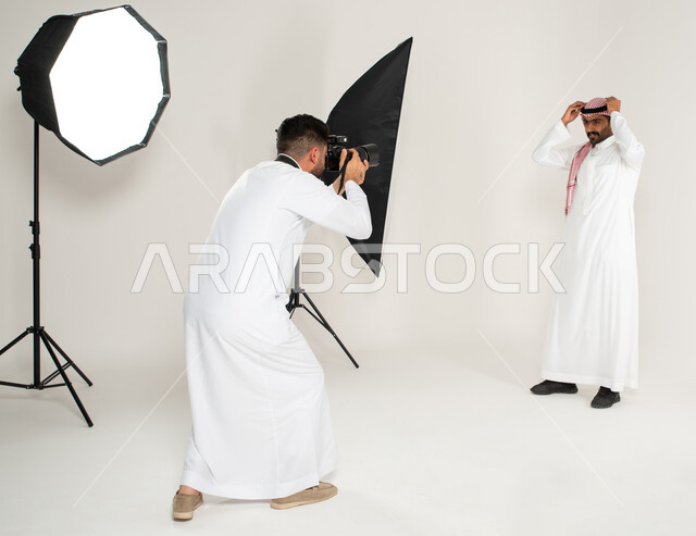 كاميرات ومعدات متطورة، بورتريه لجلسة تصوير في الاستوديو، مصور محترف عربي سعودي خليجي يقوم بالتقاط صور فوتوغرافية لرجل يرتدي الزي السعودي التقليدي، تصوير اعلاني تسويقي، خلف الكواليس، خلفية بيضاء