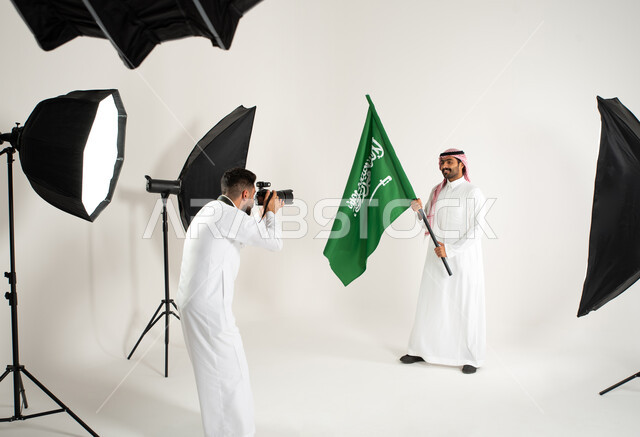 خلف الكواليس، يوم وطني سعودي، الاحتفال بيوم العلم السعودي، رجل يرتدي الزي السعودي التقليدي ويحمل عمل السعودية، مصور محترف عربي سعودي خليجي يقوم بالتقاط صور فوتوغرافية، كاميرات ومعدات متطورة، بورتريه لجلسة تصوير في الاستوديو، خلفية بيضاء
