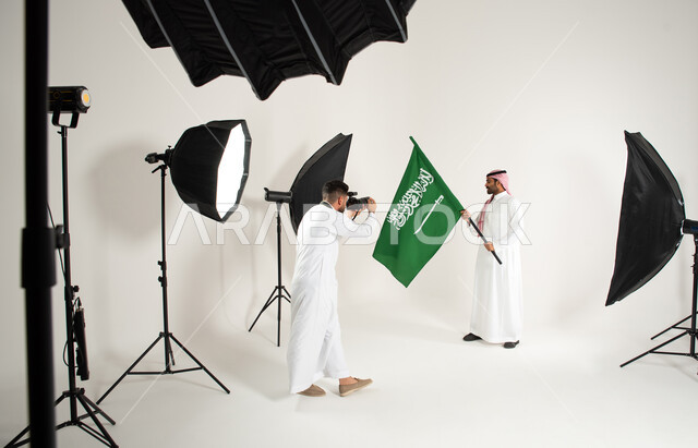 يوم وطني سعودي، رجل يرتدي الزي السعودي التقليدي ويحمل عمل السعودية، مصور محترف عربي سعودي خليجي يقوم بالتقاط صور فوتوغرافية، بورتريه لجلسة تصوير في الاستوديو، كاميرات ومعدات متطورة، خلف الكواليس، الاحتفال بيوم العلم السعودي، خلفية بيضاء