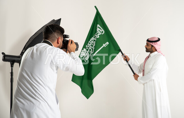 كاميرات ومعدات متطورة، خلف الكواليس، رجل يرتدي الزي السعودي التقليدي ويحمل عمل السعودية، بورتريه لجلسة تصوير في الاستوديو، مصور محترف عربي سعودي خليجي يقوم بالتقاط صور فوتوغرافية، يوم وطني سعودي، الاحتفال بيوم العلم السعودي، خلفية بيضاء