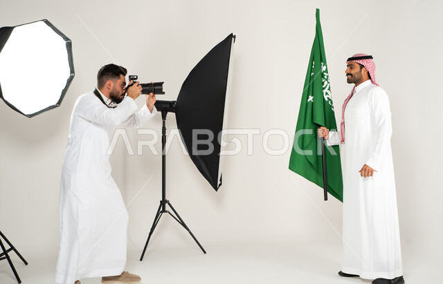 مصور محترف عربي سعودي خليجي يقوم بالتقاط صور فوتوغرافية، كاميرات ومعدات متطورة، رجل يرتدي الزي السعودي التقليدي ويحمل عمل السعودية، بورتريه لجلسة تصوير في الاستوديو، خلف الكواليس، يوم وطني سعودي، الاحتفال بيوم العلم السعودي، خلفية بيضاء