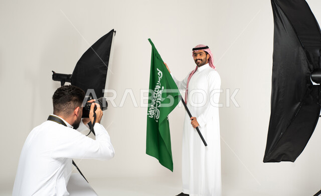 رجل يرتدي الزي السعودي التقليدي ويحمل عمل السعودية، بورتريه لجلسة تصوير في الاستوديو، مصور محترف عربي سعودي خليجي يقوم بالتقاط صور فوتوغرافية، كاميرات ومعدات متطورة، خلف الكواليس، يوم وطني سعودي، الاحتفال بيوم العلم السعودي، خلفية بيضاء