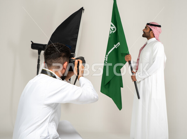 كاميرات ومعدات متطورة، يوم وطني سعودي، الاحتفال بيوم العلم السعودي، بورتريه لجلسة تصوير في الاستوديو، مصور محترف عربي سعودي خليجي يقوم بالتقاط صور فوتوغرافية لرجل يرتدي الزي السعودي التقليدي ويحمل عمل السعودية، خلف الكواليس، خلفية بيضاء