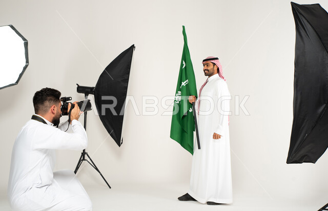 الاحتفال بيوم العلم السعودي، بورتريه لجلسة تصوير في الاستوديو، مصور محترف عربي سعودي خليجي يقوم بالتقاط صور فوتوغرافية لرجل يرتدي الزي السعودي التقليدي ويحمل عمل السعودية، كاميرات ومعدات متطورة، خلف الكواليس، يوم وطني سعودي، خلفية بيضاء