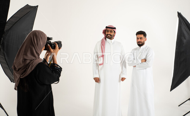 رجلان يرتديان الزي السعودي التقليدي، مصورة محترفة عربية سعودية خليجية تقوم بالتقاط صور فوتوغرافية، بورتريه لجلسة تصوير في الاستوديو، كاميرات ومعدات متطورة، خلف الكواليس، تصوير اعلاني تسويقي، خلفية بيضاء