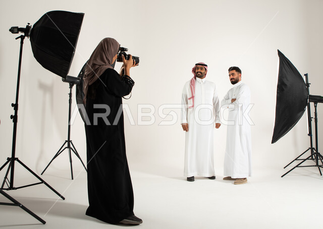 مصورة محترفة عربية سعودية خليجية تقوم بالتقاط صور فوتوغرافية، بورتريه لجلسة تصوير في الاستوديو، رجلان يرتديان الزي السعودي التقليدي، كاميرات ومعدات متطورة، خلف الكواليس، تصوير اعلاني تسويقي، خلفية بيضاء