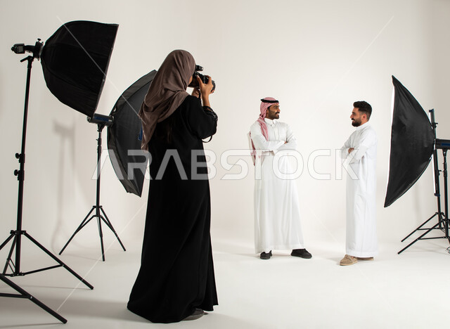 بورتريه لجلسة تصوير في الاستوديو، خلف الكواليس، تصوير اعلاني تسويقي، مصورة محترفة عربية سعودية خليجية تقوم بالتقاط صور فوتوغرافية لرجلان يرتديان الزي السعودي التقليدي، كاميرات ومعدات متطورة، خلفية بيضاء