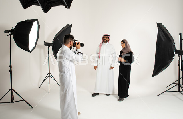 ازياء وموديلات عصرية، بورتريه لجلسة تصوير في الاستوديو، مصور محترف عربي سعودي خليجي يقوم بالتقاط صور فوتوغرافية لرجل وامرأة يرتديان الزي السعودي التقليدي، كاميرات ومعدات متطورة، تصوير اعلاني تسويقي، خلف الكواليس، خلفية بيضاء