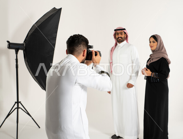 كاميرات ومعدات متطورة، أزياء وموديلات عصرية، بورتريه لجلسة تصوير في الاستوديو، مصور محترف عربي سعودي خليجي يقوم بالتقاط صور فوتوغرافية لرجل وامرأة يرتديان الزي السعودي التقليدي، تصوير اعلاني تسويقي، خلف الكواليس، خلفية بيضاء