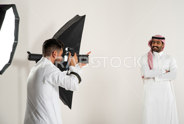 تصوير اعلاني تسويقي، بورتريه لجلسة تصوير في الاستوديو، كاميرات ومعدات متطورة، مصور محترف عربي سعودي خليجي يقوم بالتقاط صور فوتوغرافية لرجل يرتدي الزي السعودي التقليدي، خلف الكواليس، خلفية بيضاء