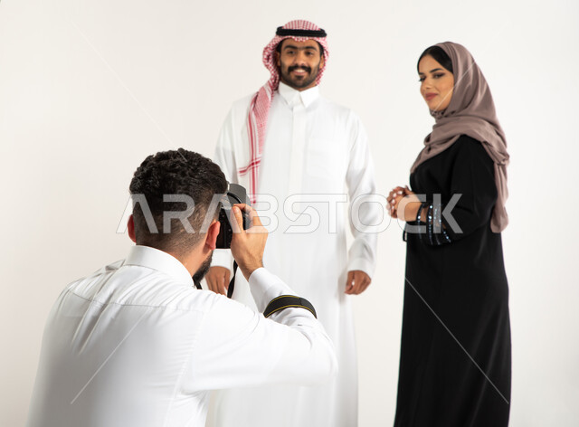 خلف الكواليس، بورتريه لجلسة تصوير في الاستوديو، مصور محترف عربي سعودي خليجي يقوم بالتقاط صور فوتوغرافية لرجل وامرأة يرتديان الزي السعودي التقليدي، كاميرات ومعدات متطورة، تصوير اعلاني تسويقي، أزياء وموديلات عصرية، خلفية بيضاء