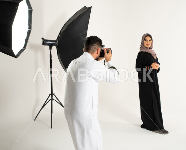 عارضة أزياء وموديلات عصرية، بورتريه لجلسة تصوير في الاستوديو، مصور محترف عربي سعودي خليجي يقوم بالتقاط صور فوتوغرافية لامرأة ترتدي عباءة نسائية خليجية، كاميرات ومعدات متطورة، تصوير اعلاني تسويقي، خلف الكواليس، خلفية بيضاء