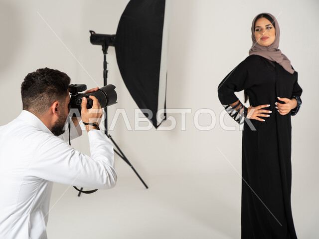 مصور محترف عربي سعودي خليجي يقوم بالتقاط صور فوتوغرافية لامرأة ترتدي عباءة نسائية خليجية، بورتريه لجلسة تصوير في الاستوديو، كاميرات ومعدات متطورة، تصوير اعلاني تسويقي، خلف الكواليس، عارضة أزياء وموديلات عصرية، خلفية بيضاء