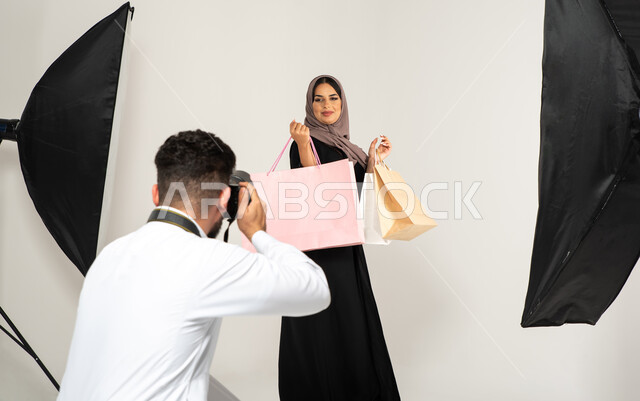 تصوير اعلاني تسويقي، بورتريه لجلسة تصوير في الاستوديو، مصور محترف عربي سعودي خليجي يقوم بالتقاط صور فوتوغرافية لامرأة تحمل بيدها اكياس التسوق، كاميرات ومعدات متطورة، عارضة أزياء وموديلات عصرية، خلفية بيضاء، خلف الكواليس