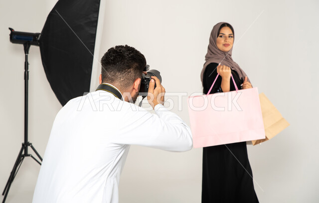 كاميرات ومعدات متطورة، بورتريه لجلسة تصوير في الاستوديو، مصور محترف عربي سعودي خليجي يقوم بالتقاط صور فوتوغرافية لامرأة تحمل بيدها اكياس التسوق، تصوير اعلاني تسويقي، عارضة أزياء وموديلات عصرية، خلفية بيضاء، خلف الكواليس
