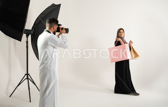 عارضة أزياء وموديلات عصرية، بورتريه لجلسة تصوير في الاستوديو، مصور محترف عربي سعودي خليجي يقوم بالتقاط صور فوتوغرافية لامرأة تحمل بيدها اكياس التسوق، تصوير اعلاني تسويقي، كاميرات ومعدات متطورة، خلفية بيضاء، خلف الكواليس