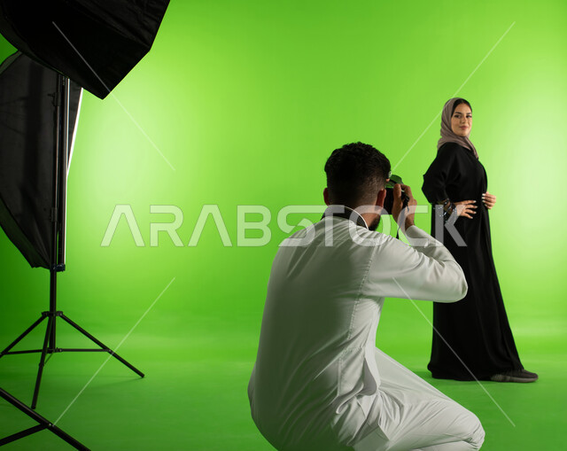 تصوير اعلاني تسويقي، كروما لجلسة تصوير في الاستوديو، مصور محترف عربي سعودي خليجي يقوم بالتقاط صور فوتوغرافية لامرأة ترتدي عباءة نسائية خليجية، كاميرات ومعدات متطورة، عارضة أزياء وموديلات عصرية، خلفية شاشة خضراء، خلف الكواليس