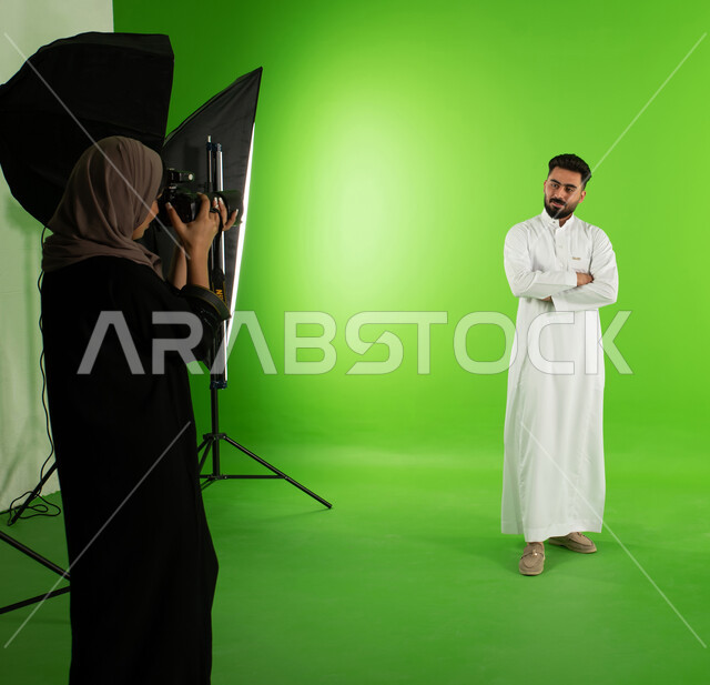 مصورة محترفة عربية سعودية خليجية تقوم بالتقاط صور فوتوغرافية لرجل يقف بايدي مكتوفة ومتقاطعة، كروما لجلسة تصوير في الاستوديو، تصوير اعلاني تسويقي، كاميرات ومعدات متطورة، خلفية شاشة خضراء، خلف الكواليس