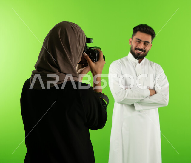 مصورة محترفة عربية سعودية خليجية تقوم بالتقاط صور فوتوغرافية لرجل يقف بايدي مكتوفة ومتقاطعة، كروما لجلسة تصوير في الاستوديو، تصوير اعلاني تسويقي، كاميرات ومعدات متطورة، خلفية شاشة خضراء، خلف الكواليس
