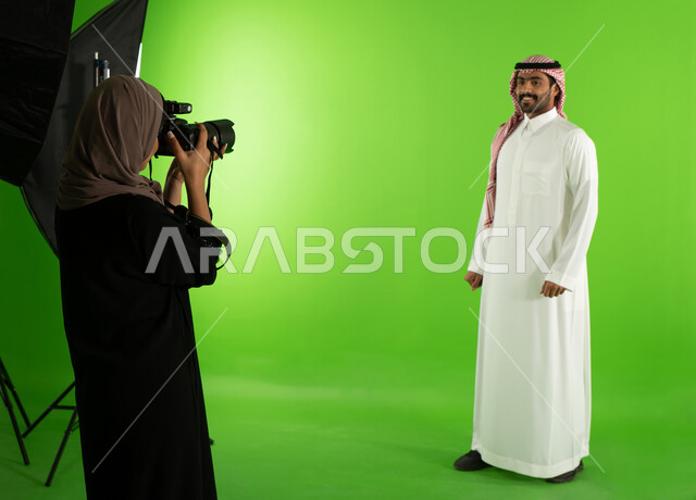 كروما لجلسة تصوير في الاستوديو، رجل يرتدي الزي السعودي التقليدي، مصورة محترفة عربية سعودية خليجية تقوم بالتقاط صور فوتوغرافية، تصوير اعلاني تسويقي، كاميرات ومعدات متطورة، خلفية شاشة خضراء، خلف الكواليس