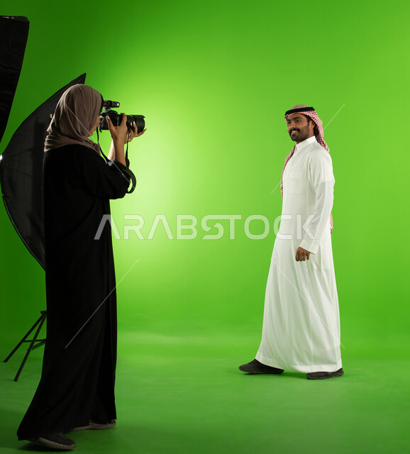 تصوير اعلاني تسويقي، مصورة محترفة عربية سعودية خليجية تقوم بالتقاط صور فوتوغرافية لرجل يرتدي الزي السعودي التقليدي، كروما لجلسة تصوير في الاستوديو، كاميرات ومعدات متطورة، خلفية شاشة خضراء، خلف الكواليس