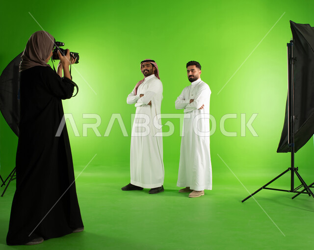 تصوير اعلاني تسويقي، مصورة محترفة عربية سعودية خليجية تقوم بالتقاط صور فوتوغرافية لرجلان يرتديان الزي السعودي التقليدي، كاميرات ومعدات متطورة، كروما لجلسة تصوير في الاستوديو، خلفية شاشة خضراء، خلف الكواليس