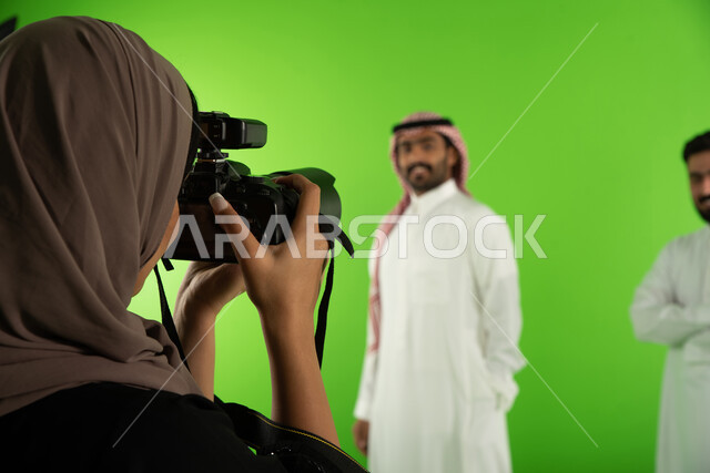صورة مقربة لمصورة محترفة عربية سعودية خليجية تقوم بالتقاط صور فوتوغرافية لرجلان يرتديان الزي السعودي التقليدي، كاميرات ومعدات متطورة، تصوير اعلاني تسويقي، كروما لجلسة تصوير في الاستوديو، خلفية شاشة خضراء، خلف الكواليس