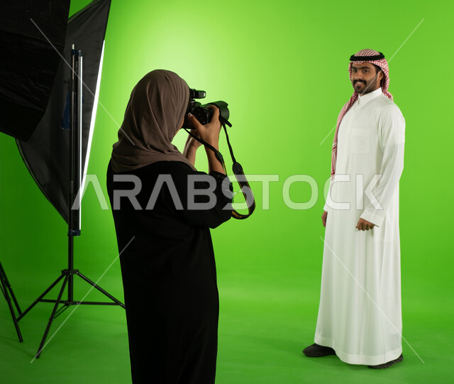 مصورة محترفة عربية سعودية خليجية تقوم بالتقاط صور فوتوغرافية لرجل يرتدي الزي السعودي التقليدي، كروما لجلسة تصوير في الاستوديو، كاميرات ومعدات متطورة، تصوير اعلاني تسويقي، خلفية شاشة خضراء، خلف الكواليس
