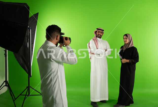 تصوير اعلاني تسويقي، كروما لجلسة تصوير في الاستوديو، مصور محترف عربي سعودي خليجي يقوم بالتقاط صور فوتوغرافية لرجل وامرأة يرتديان الزي السعودي التقليدي، كاميرات ومعدات متطورة، أزياء وموديلات عصرية، خلفية شاشة خضراء، خلف الكواليس