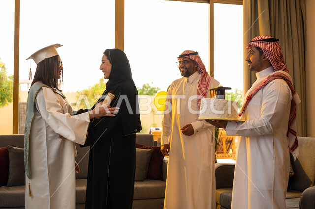 A mother celebrates her daughter's graduation with success and excellence, a happy Saudi Gulf Arab family, university graduation celebration, cake mold decorated with a graduation cap, a university graduate wearing graduation clothes and holding a certifi