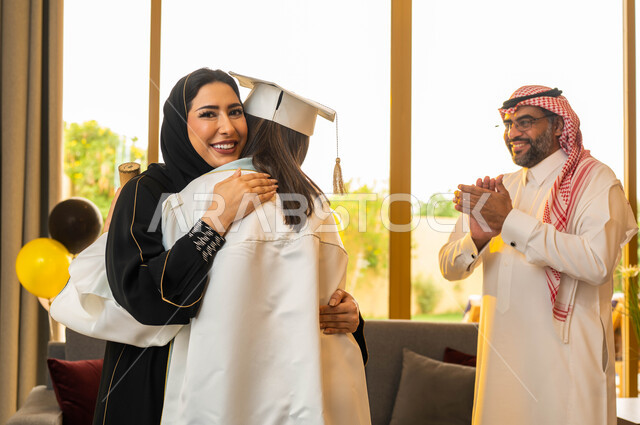 A happy Saudi Gulf Arab family celebrates their daughter's graduation from the university. A university graduate wears graduation clothes and holds a certificate of excellence. A mother hugs her daughter with joy at her graduation from the university with