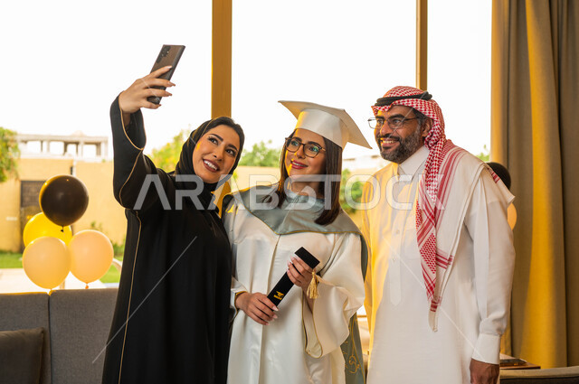 A university graduate wearing graduation clothes and holding a certificate of success, taking commemorative selfies using a mobile phone, wearing medical glasses, facial gestures indicating happiness, a happy Saudi Gulf Arab family, university graduation 
