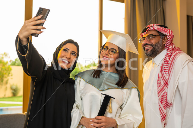 Wearing medical glasses, taking memorial selfies using a mobile phone, a university graduate wearing graduation clothes and holding a certificate of success, facial gestures indicating happiness, a happy Saudi Gulf Arab family, university graduation celeb