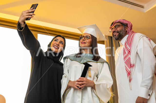 التقاط صور سيلفي تذكارية باستخدام الهاتف المحمول، خريجة جامعية ترتدي ملابس التخرج وتمسك شهادة النجاح، ارتداء النظارات الطبية، ايماءات وجه تدل على السعادة، عائلة سعودية خليجية عربية سعيدة، الاحتفال بالتخرج من الجامعة، التعليم في السعودية