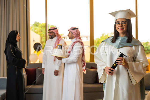 عائلة سعودية خليجية عربية تحتفل بتخرج ابنتها من الجامعة، قالب الجاتوه المزين بقبعة التخرج، خريجة جامعية ترتدي النظارات الطبية وملابس التخرج وتمسك شهادة النجاح والتفوق، ايماءات وجه تدل على السعادة، التعليم في السعودية
