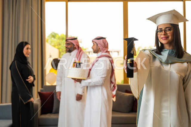 خريجة جامعية ترتدي النظارات الطبية وملابس التخرج وتمسك شهادة النجاح والتفوق، قالب الجاتوه المزين بقبعة التخرج، ايماءات وجه تدل على السعادة، عائلة سعودية خليجية عربية تحتفل بتخرج ابنتها من الجامعة، التعليم في السعودية