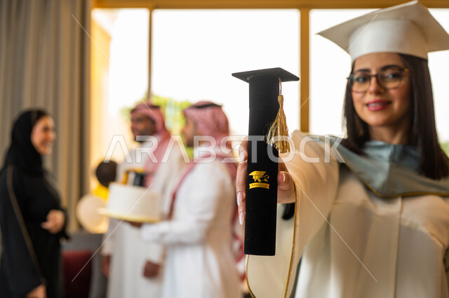 قالب الجاتوه المزين بقبعة التخرج، خريجة جامعية ترتدي النظارات الطبية وملابس التخرج وتمسك شهادة النجاح والتفوق، ايماءات وجه تدل على السعادة، عائلة سعودية خليجية عربية تحتفل بتخرج ابنتها من الجامعة، التعليم في السعودية