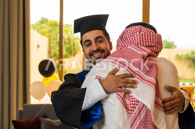 Celebrating university graduation, a Saudi Arabian Gulf man hugging his son with joy at his graduation from the university with excellence and distinction, a university graduate wearing graduation clothes and holding a certificate of excellence and succes