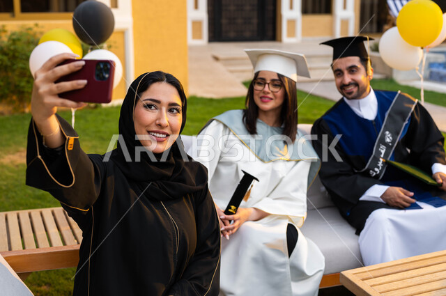 الاحتفال بالتخرج من الجامعة، ام تقوم بالتقاط الصور التذكارية من خلال الهاتف المحمول، عائلة سعودية خليجية عربية سعيدة، خريج وخريجة جامعيين يرتديان ملابس التخرج ويمسكان شهادة النجاح والتفوق، ايماءات وجه تدل على السعادة، التعليم في السعودية