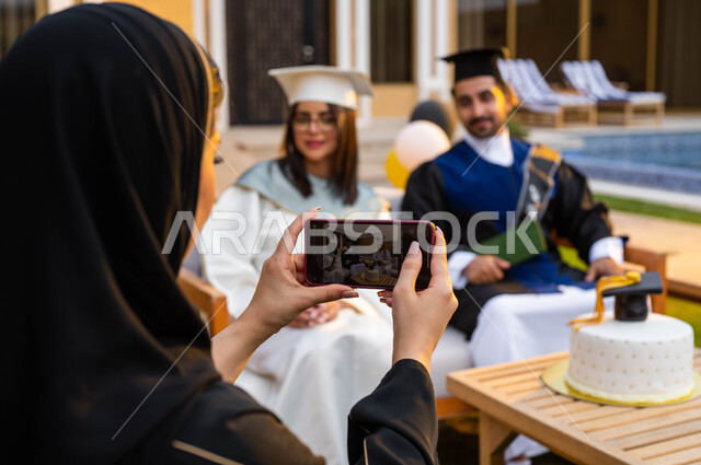 ام تقوم بالتقاط الصور التذكارية لابنائها من خلال الهاتف المحمول، كيكة مزينة بقبعة التخرج، الاحتفال بالتخرج من الجامعة، خريج وخريجة جامعيين يرتديان ملابس التخرج ويمسكان شهادة النجاح والتفوق، التعليم في السعودية