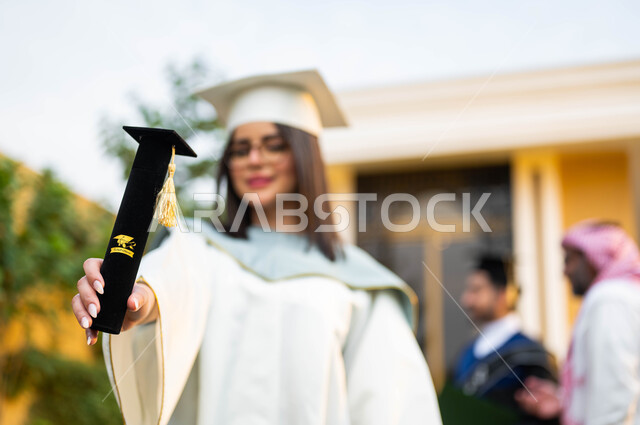 خريجة جامعية بايماءات وجه تدل على السعادة والفرح، شابة عربية خليجية سعودية ترتدي ملابس التخرج وتمسك شهادة النجاح والتفوق، عائلة سعودية خليجية عربية سعيدة تحتفل بتخرج ابنائها من الجامعة، التعليم في السعودية