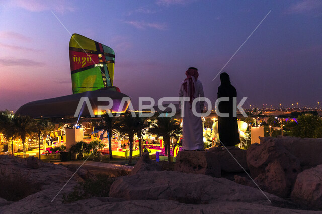 منارة ثقافية و معالم سياحية مشهورة، صورة من الخلف لرجل و امرأة يرتديان الزي السعودي يقفان على الحجارة بالقرب من مركز الملك عبدالعزيز الثقافي العالمي في مدينة الظهران في المملكة العربية السعودية، موسم تنوين للأنشطة الترفيهية و التعليمية و الثقافية .