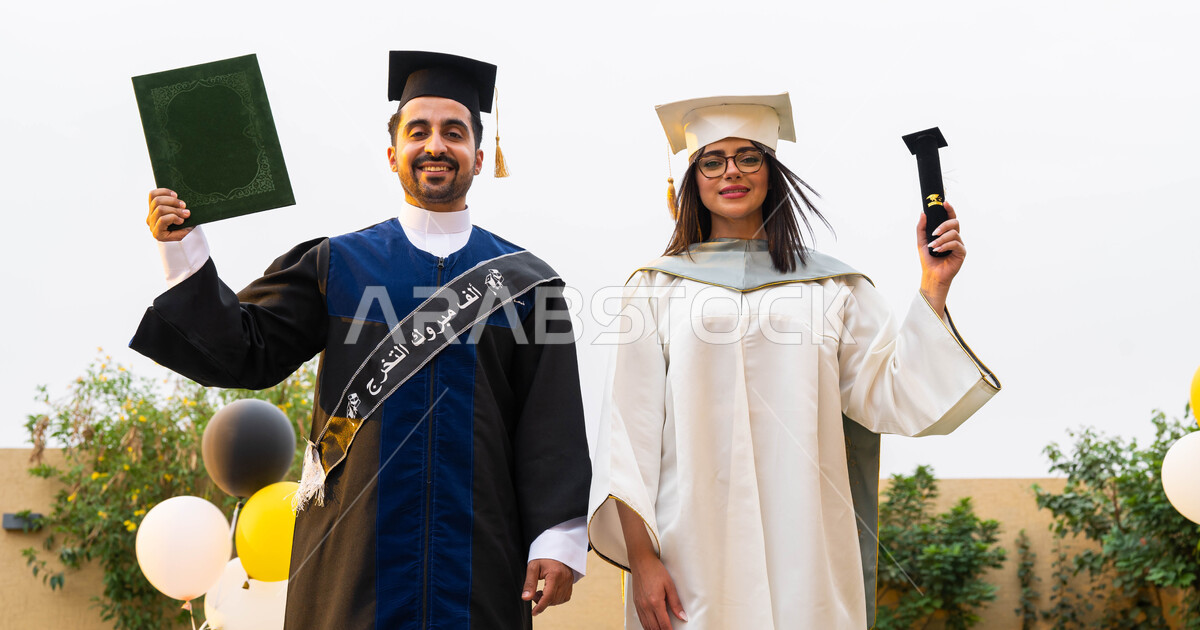 خريج وخريجة جامعيين سعوديان خليجيان عربيان يرتديان ملابس التخرج ويمسكان ...